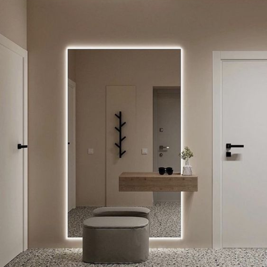 Backlit rectangular mirror above a floating wooden console; two round gray ottomans in front with a small plant and sunglasses on the console, terrazzo floor in a beige entryway.