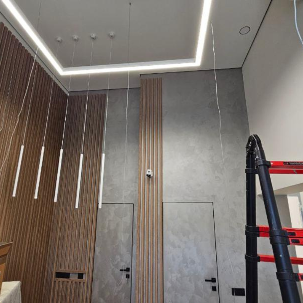 Modern interior under construction with a wooden slat wall on the left, gray concrete walls, and hanging light pendants alongside a ceiling LED frame.