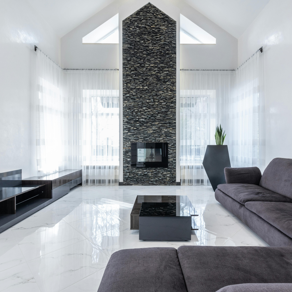 Modern living room with a tall stacked-stone fireplace, white walls, and sheer curtains flanking large windows.