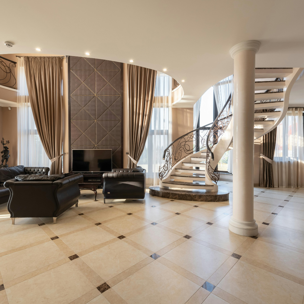 Luxurious living room with curved staircase, black leather seating, marble floor, and tall beige curtains.