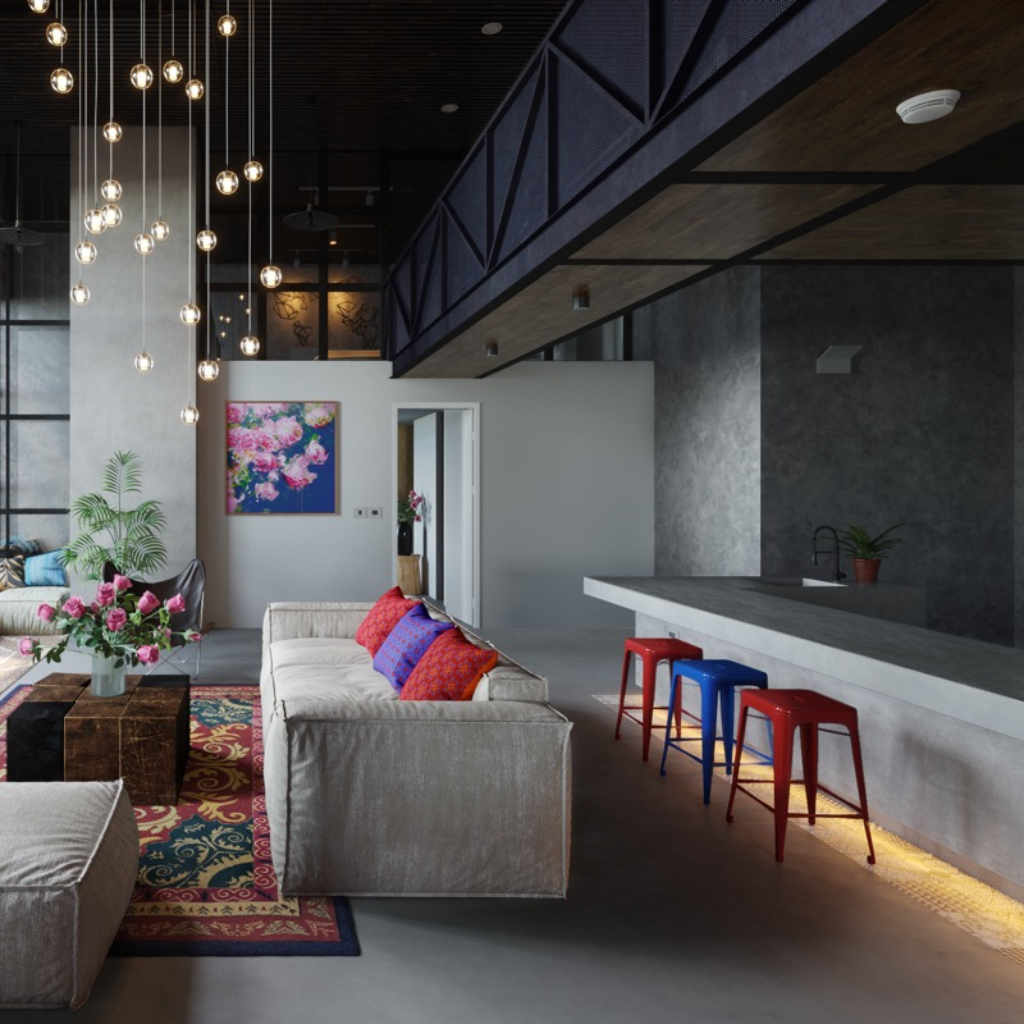 Open lounge with a gray sectional, multicolored cushions, a vibrant rug, and a concrete bar with red and blue stools under hanging bulbs.