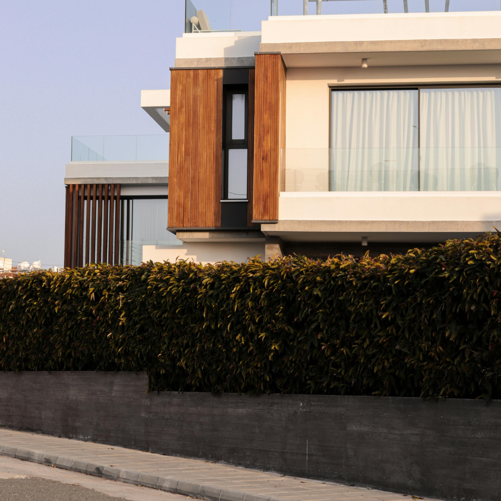 Modern two-story building with vertical wooden panels, glass balcony, and white facade, set behind a tall hedge on a concrete wall.