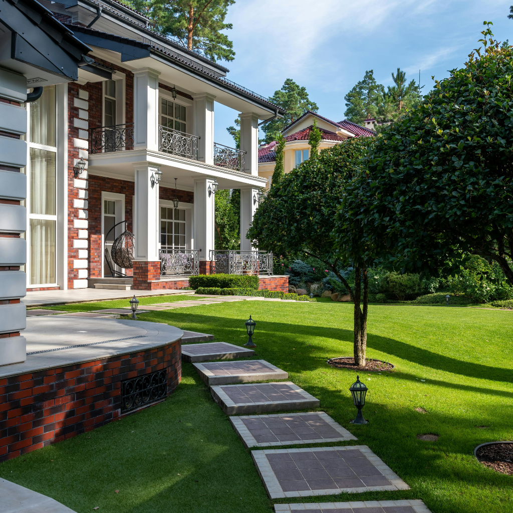 Front yard of a brick-and-white two-story house with tall white columns, ornate iron balconies, and a stepping-stone path across a green lawn.
