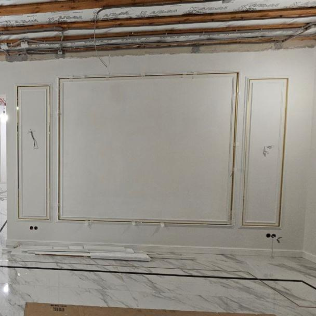 Under-construction living room with a large white paneled wall featuring gold trim, exposed ceiling wires, and a marble floor outside the finished area.