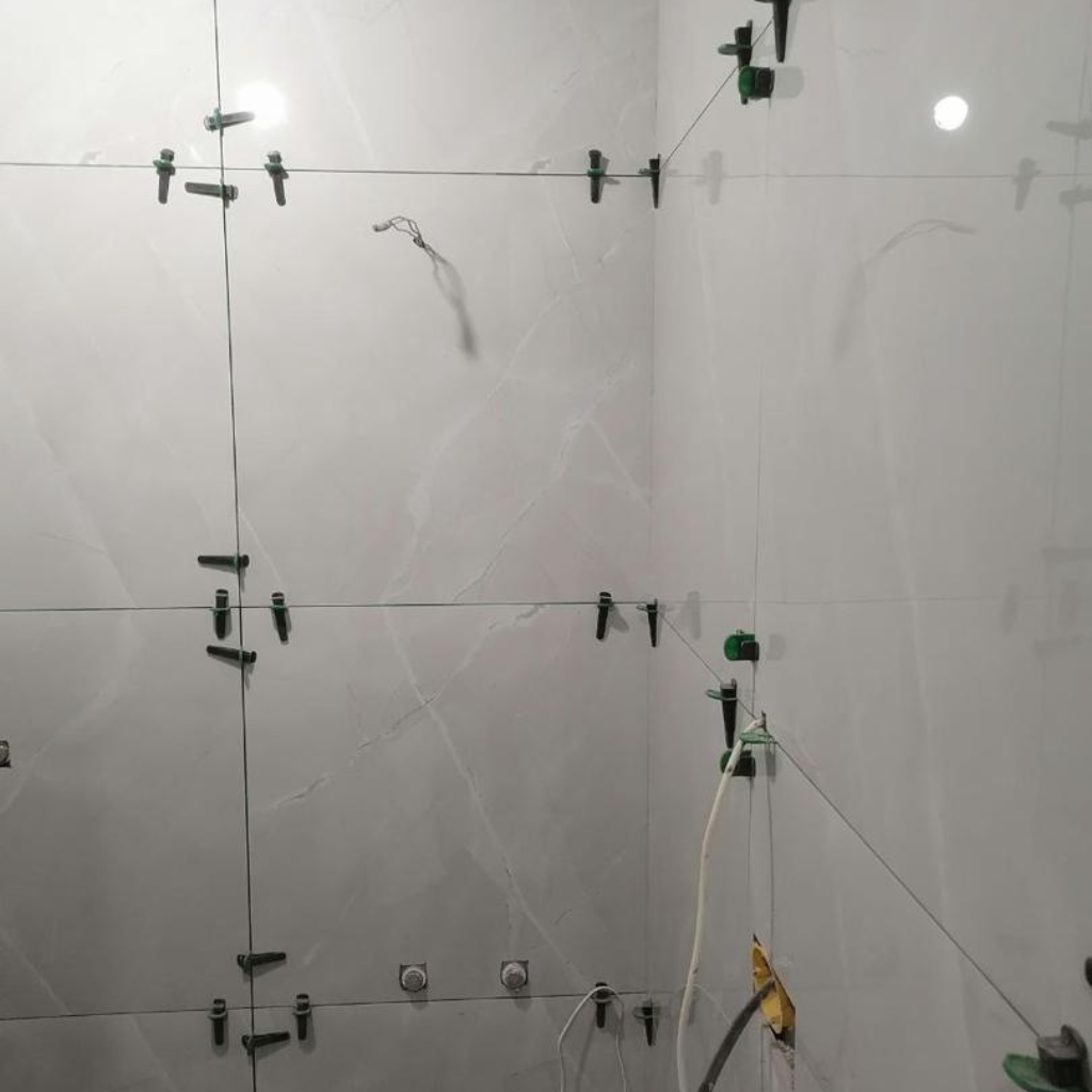 Tiled wall with green tile spacers and clips during installation; a white hose hangs on the right side.