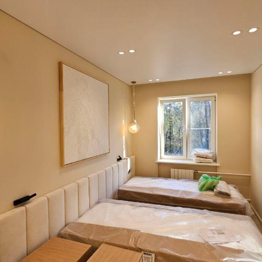 Hotel room with two single beds and a padded headboard, beige walls, a large abstract canvas, and a window overlooking trees.