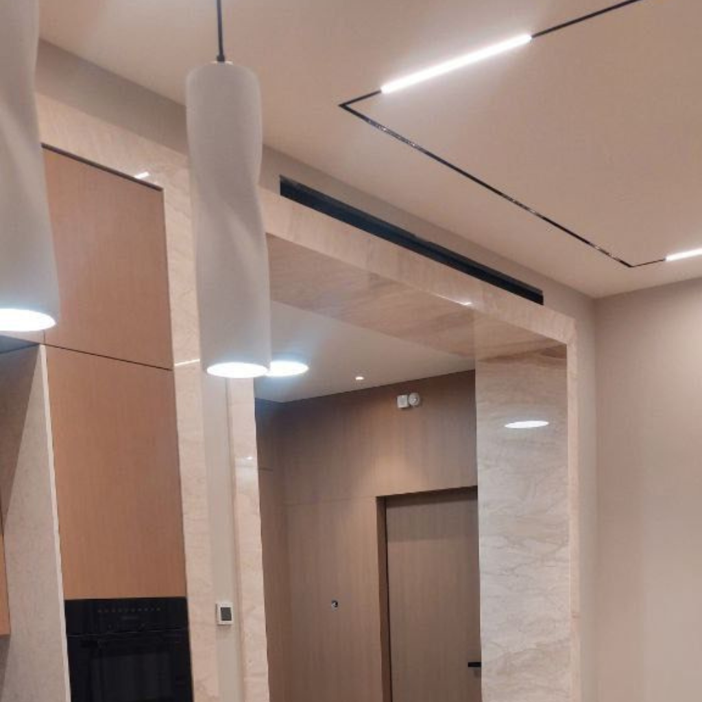 Modern indoor lobby area with beige walls, marble columns, wooden panels, and cylindrical pendant lights overhead.