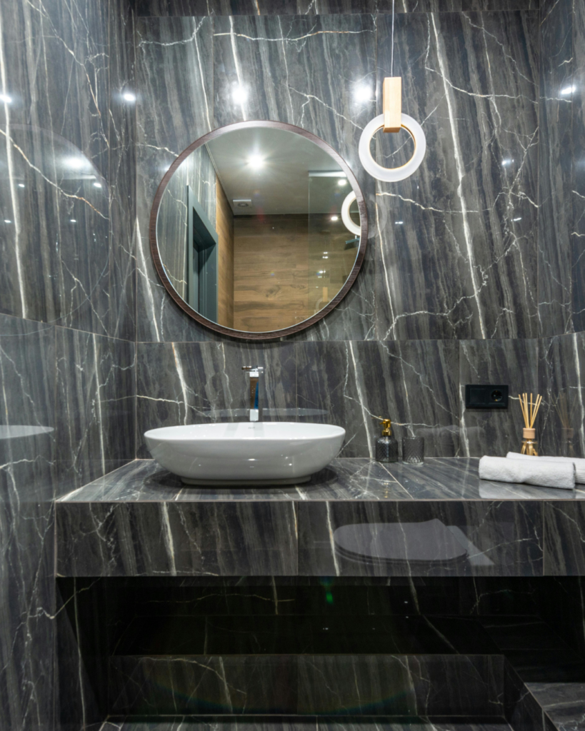 Round mirror above a white vessel sink on a dark marble vanity with a towel ring and accessories to the right in a luxurious bathroom visually dominated by marble.