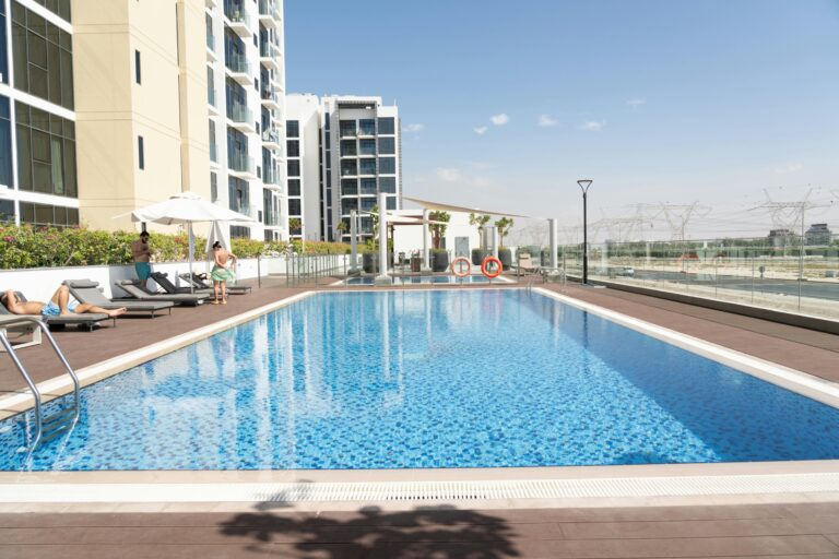 Outdoor pool deck at a modern high-rise apartment complex, with lounge chairs, umbrellas, and residents relaxing along the pool edge under a blue sky.