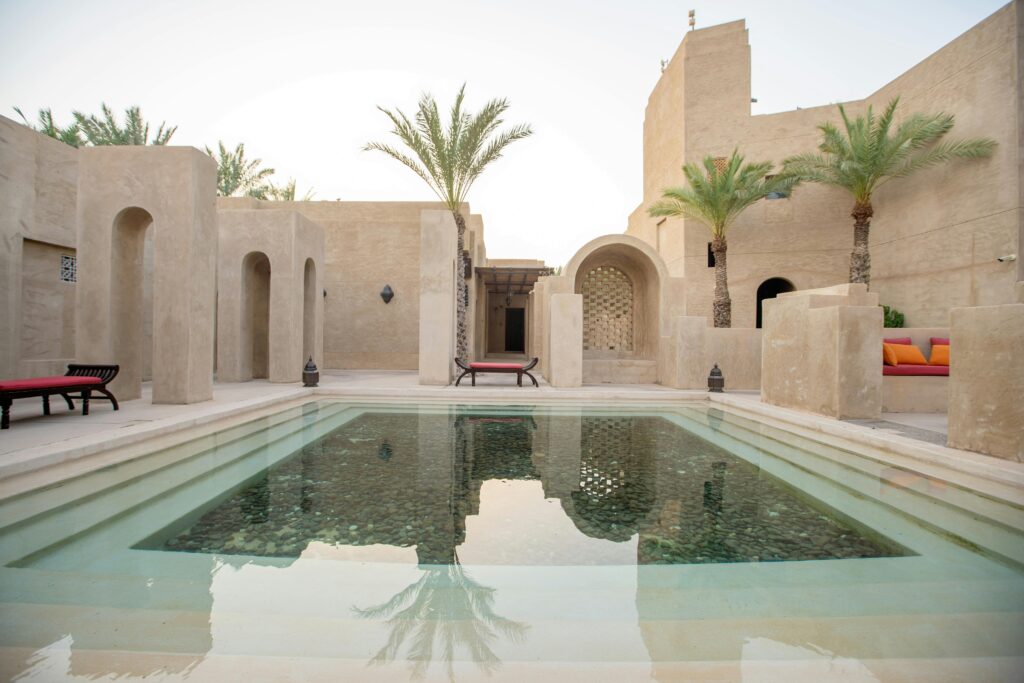 Sunlit desert courtyard with beige stucco walls, palm trees, arches, and a rectangular shallow pool reflecting the sky.