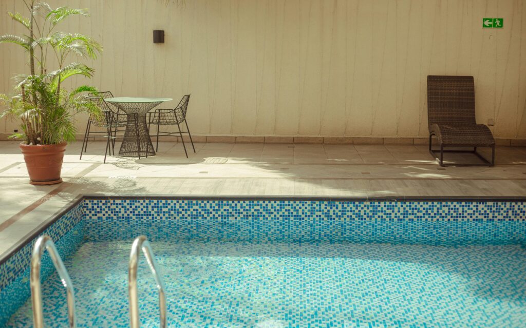 Indoor pool area with blue mosaic tiles, a metal ladder, and wicker seating including a glass table and chairs beside a potted plant on a beige wall backdrop.